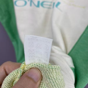 Puede incluir: Primer plano de una sudadera blanca y verde con el nombre de la marca "O'NEIL" en turquesa. Se ve una etiqueta de cuidado, con s&iacute;mbolos que indican las instrucciones de lavado. El pu&ntilde;o es de color verde claro y amarillo.