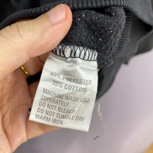 May include: Close-up of a black garment's tag. The tag reads "65% POLYESTER 35% COTTON" and washing instructions: "MACHINE WASH COLD SEPERATELY DO NOT BLEACH DO NOT TUMBLE DRY WARM IRON". The garment's fabric is visible.