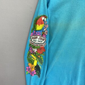 K&ouml;nnte beinhalten: Ein helles t&uuml;rkisfarbenes Langarmshirt mit einem farbenfrohen Grafikdruck auf dem &Auml;rmel. Das Design zeigt einen Papagei, einen Tukan, tropische Blumen, einen Schmetterling und den Text "RON JON SURF SHOP ONE AND ONLY" in einem wei&szlig;en Oval.