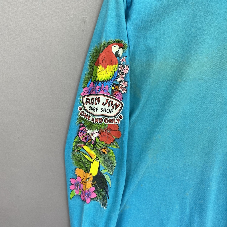 K&ouml;nnte beinhalten: Ein helles t&uuml;rkisfarbenes Langarmshirt mit einem farbenfrohen Grafikdruck auf dem &Auml;rmel. Das Design zeigt einen Papagei, einen Tukan, tropische Blumen, einen Schmetterling und den Text "RON JON SURF SHOP ONE AND ONLY" in einem wei&szlig;en Oval.