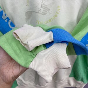 Puede incluir: Primer plano de una sudadera con bloques de color con detalles en verde, azul y blanco. Las mangas tienen pu&ntilde;os acanalados en blanco. La sudadera tiene el texto "THE SPORTSWEAR" y un dise&ntilde;o gr&aacute;fico.