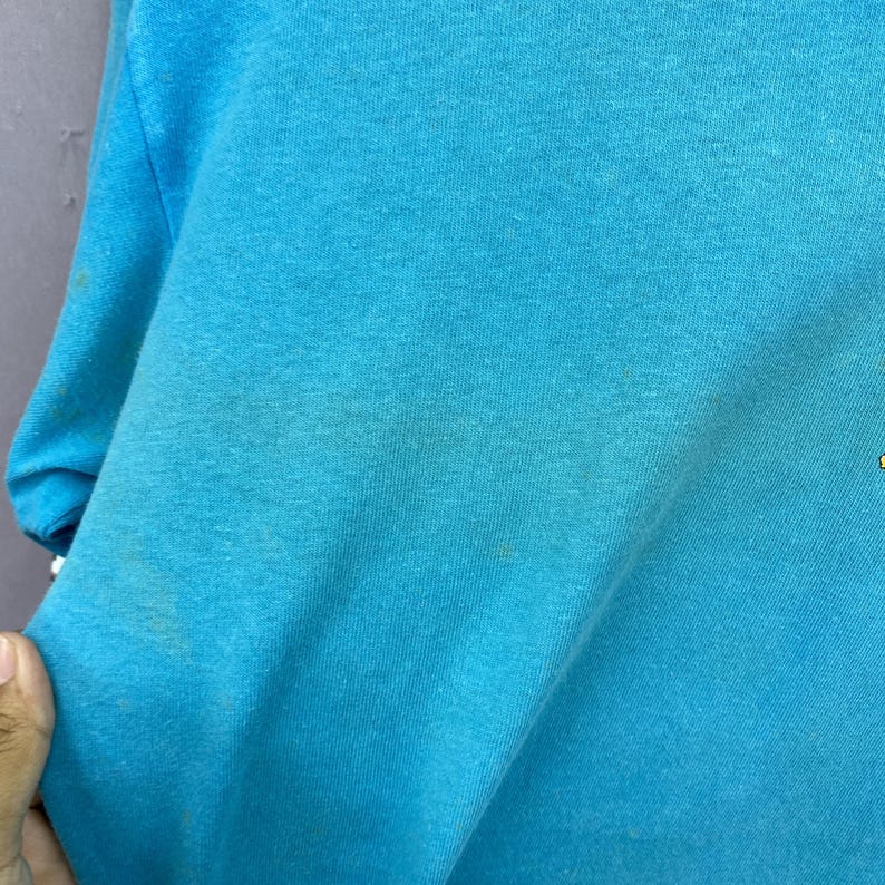 K&ouml;nnte beinhalten: Ein leuchtend t&uuml;rkisfarbenes T-Shirt aus weichem Stoff. Das Kleidungsst&uuml;ck weist eine durchgehend kr&auml;ftige, einfarbige Optik auf dem sichtbaren Teil des Materials auf, mit einer glatten Textur und einer klassischen Kurzarmkonstruktion auf der linken Seite.