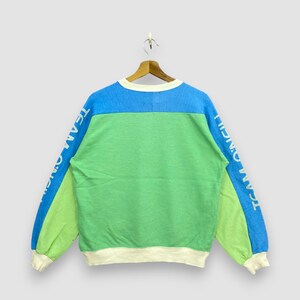 Puede incluir: Una sudadera con bloques de color en azul, verde y verde claro, con el texto "TEAM O'NEILL" impreso verticalmente en las mangas. La sudadera tiene cuello redondo y pu&ntilde;os y dobladillo acanalados.