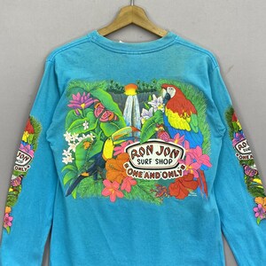 K&ouml;nnte beinhalten: Ein helles, t&uuml;rkisfarbenes Langarmshirt mit einer lebendigen tropischen Grafik auf der R&uuml;ckseite. Das Design zeigt einen Papagei, einen Tukan, einen Wasserfall und verschiedene Blumen. Der Text lautet RON JON SURF SHOP ONE AND ONLY.