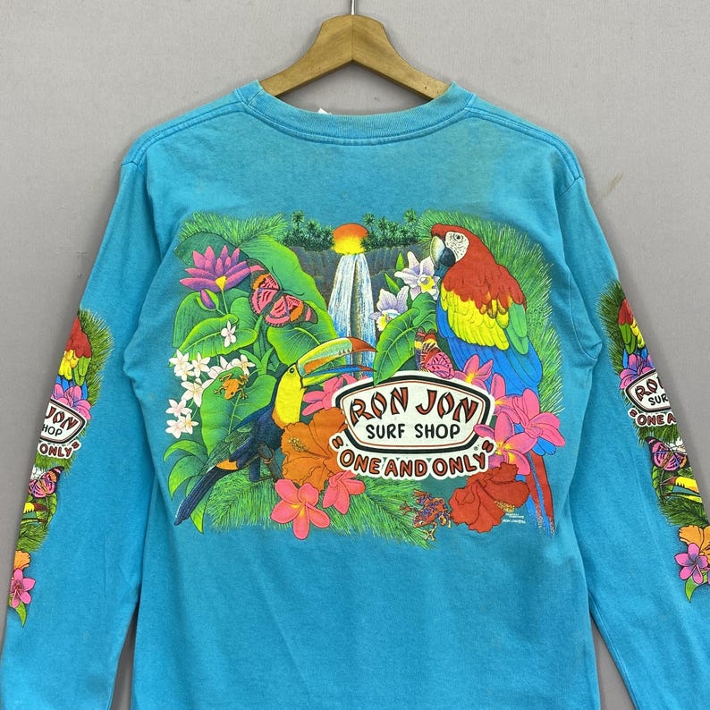 K&ouml;nnte beinhalten: Ein helles, t&uuml;rkisfarbenes Langarmshirt mit einer lebendigen tropischen Grafik auf der R&uuml;ckseite. Das Design zeigt einen Papagei, einen Tukan, einen Wasserfall und verschiedene Blumen. Der Text lautet RON JON SURF SHOP ONE AND ONLY.