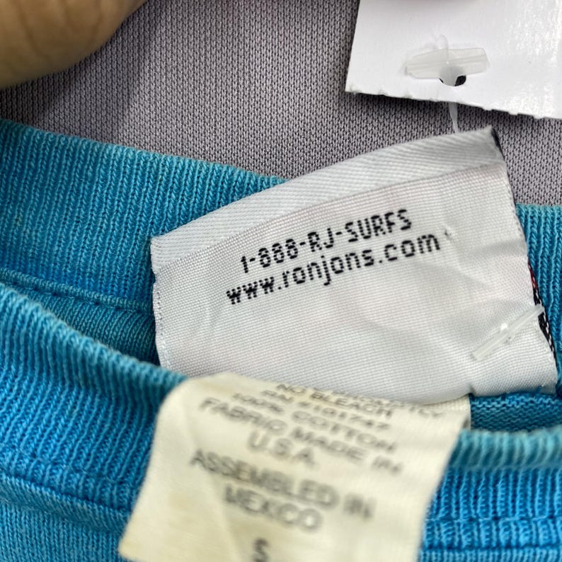 K&ouml;nnte beinhalten: Ein helles t&uuml;rkisfarbenes T-Shirt mit einem wei&szlig;en Innenetikett mit dem Text 1-888-RJ-SURFS und www.ronjons.com. Ein zweites wei&szlig;es Etikett darunter zeigt Text, einschlie&szlig;lich 100% COTTON, FABRIC MADE IN U.S.A., ASSEMBLED IN MEXICO und die Gr&ouml;&szlig;e S.