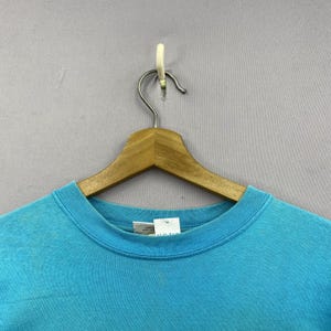 K&ouml;nnte beinhalten: Ein helles, t&uuml;rkisfarbenes T-Shirt mit Rundhalsausschnitt aus Baumwollstoff. Das Shirt verf&uuml;gt &uuml;ber einen gerippten Kragen und ein wei&szlig;es Innenetikett am Nacken mit sichtbarem Text. Es h&auml;ngt an einem Holzkleiderb&uuml;gel vor einem grauen Hintergrund.