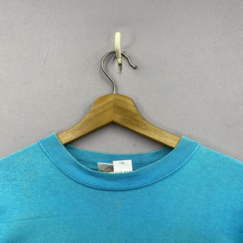 K&ouml;nnte beinhalten: Ein helles, t&uuml;rkisfarbenes T-Shirt mit Rundhalsausschnitt aus Baumwollstoff. Das Shirt verf&uuml;gt &uuml;ber einen gerippten Kragen und ein wei&szlig;es Innenetikett am Nacken mit sichtbarem Text. Es h&auml;ngt an einem Holzkleiderb&uuml;gel vor einem grauen Hintergrund.