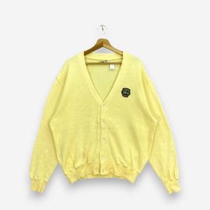 May include: A light yellow cardigan sweater with a V-neck and button closure. The sweater features a small emblem on the chest. The sweater is hanging on a wooden hanger.