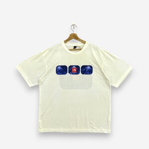 Puede incluir: Una camiseta de manga corta de color crema con un estampado gráfico en el pecho. El gráfico consta de tres marcos rectangulares redondeados con imágenes de montañas azules y un cuadrado rojo con el logotipo de The North Face en el centro.