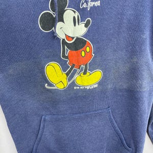 K&ouml;nnte beinhalten: Ein marineblauer Kapuzenpullover mit einer Mickey Mouse-Grafik. Die Cartoon-Maus tr&auml;gt rote Shorts und gelbe Schuhe. Das Wort "California" steht &uuml;ber der Figur. Der Kapuzenpullover hat eine Vordertasche.