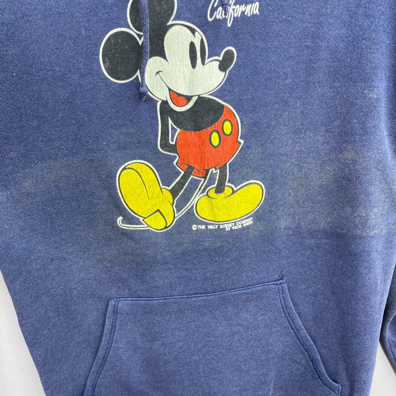 K&ouml;nnte beinhalten: Ein marineblauer Kapuzenpullover mit einer Mickey Mouse-Grafik. Die Cartoon-Maus tr&auml;gt rote Shorts und gelbe Schuhe. Das Wort "California" steht &uuml;ber der Figur. Der Kapuzenpullover hat eine Vordertasche.