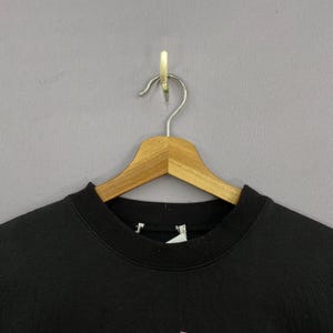 May include: A black sweatshirt hangs on a wooden hanger against a gray background. The sweatshirt has a crew neck and a tag is visible. The bottom of the sweatshirt has a pink design.
