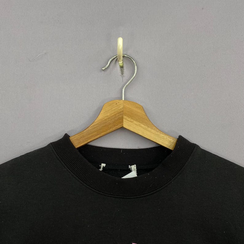 May include: A black sweatshirt hangs on a wooden hanger against a gray background. The sweatshirt has a crew neck and a tag is visible. The bottom of the sweatshirt has a pink design.