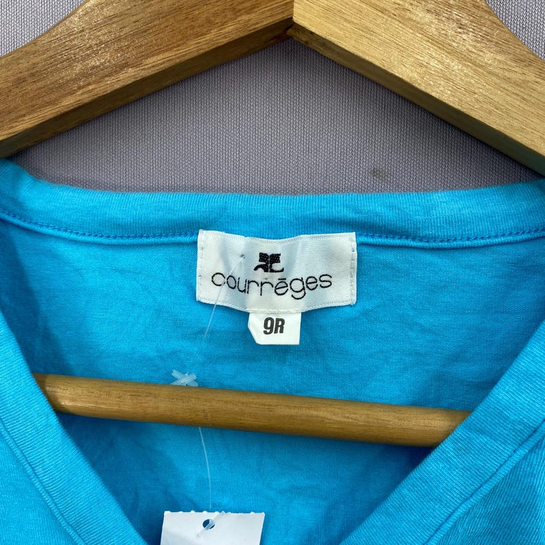 以下が含まれることがあります： ターコイズブルーのCourr&egrave;gesのVネックTシャツ。白いラベルにはブランド名とサイズ9Rが表示されています。シャツは木製のハンガーにかかっています。