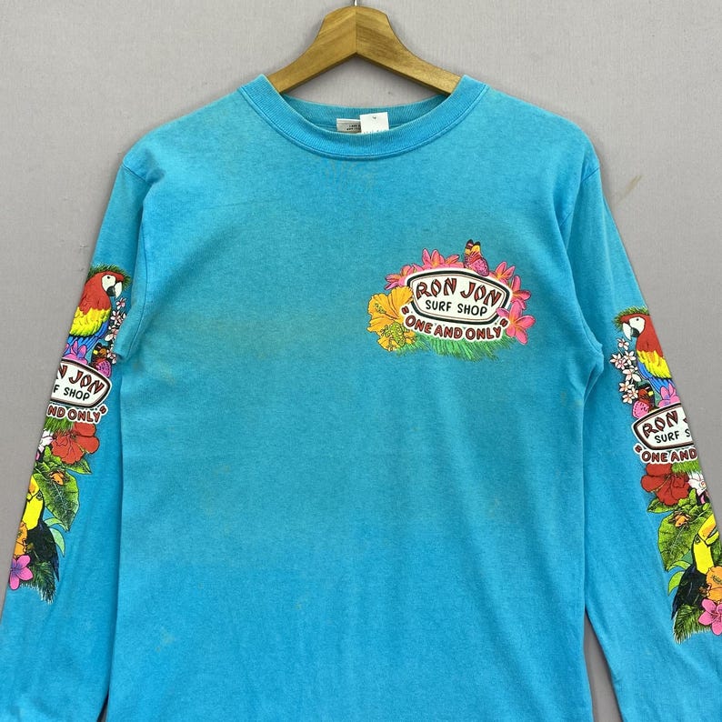 K&ouml;nnte beinhalten: Ein hellt&uuml;rkisfarbenes Langarmshirt mit Rundhalsausschnitt und einem Grafikdruck auf der Brust sowie beiden &Auml;rmeln. Die Brustgrafik zeigt den Text "RON JON SURF SHOP ONE AND ONLY", umgeben von tropischen Blumen, einem Papagei und einem Tukan.