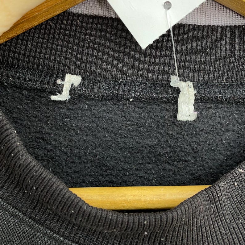 May include: Close-up of a black sweatshirt with a tag attached. The sweatshirt has a ribbed neckline and a soft, fleecy interior. The tag is white and appears to be made of paper. The garment is hanging on a wooden hanger.