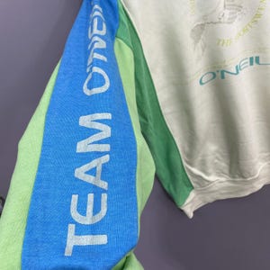 Puede incluir: Una sudadera con bloques de color verde claro, azul y blanco roto. La manga izquierda presenta el texto "TEAM O'NEILL" en blanco. La sudadera tiene cuello redondo y un gr&aacute;fico en la parte superior derecha del pecho.