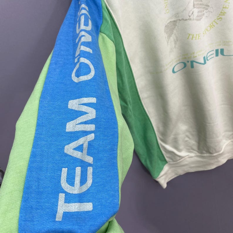 Puede incluir: Una sudadera con bloques de color verde claro, azul y blanco roto. La manga izquierda presenta el texto "TEAM O'NEILL" en blanco. La sudadera tiene cuello redondo y un gr&aacute;fico en la parte superior derecha del pecho.