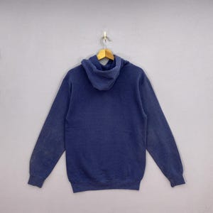 K&ouml;nnte beinhalten: Ein marineblauer Kapuzenpullover mit Kordelzug, langen &Auml;rmeln und einer K&auml;ngurutasche. Der Pullover h&auml;ngt an einem Holzb&uuml;gel vor grauem Hintergrund. Der Stoff scheint weich und abgenutzt zu sein.
