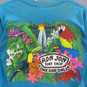 K&ouml;nnte beinhalten: Ein t&uuml;rkisfarbenes Langarmshirt mit einer tropischen Grafik auf der R&uuml;ckseite. Das Motiv zeigt einen Wasserfall, einen Tukan, einen Ara, Schmetterlinge, Fr&ouml;sche und bunte Blumen. Der Text lautet RON JON SURF SHOP ONE AND ONLY.