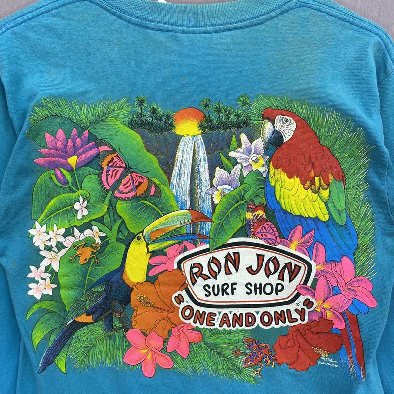 K&ouml;nnte beinhalten: Ein t&uuml;rkisfarbenes Langarmshirt mit einer tropischen Grafik auf der R&uuml;ckseite. Das Motiv zeigt einen Wasserfall, einen Tukan, einen Ara, Schmetterlinge, Fr&ouml;sche und bunte Blumen. Der Text lautet RON JON SURF SHOP ONE AND ONLY.