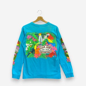 K&ouml;nnte beinhalten: Ein hellt&uuml;rkisfarbenes Langarmshirt mit einer bunten Grafik auf der R&uuml;ckseite, die einen Tukan, einen Ara, tropische Blumen und einen Wasserfall zeigt. Der Text lautet Ron Jon Surf Shop One and Only. &Auml;hnliche tropische Motive sind auf den &Auml;rmeln.