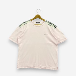 Könnte beinhalten: Hellrosa T-Shirt mit kurzen Ärmeln, an einem Holzbügel hängend. Die Schultern haben ein schwarz-weiß-grünes Karomuster. Auf der Brust des Shirts steht der Text "BURBERRY BLACK LABEL".