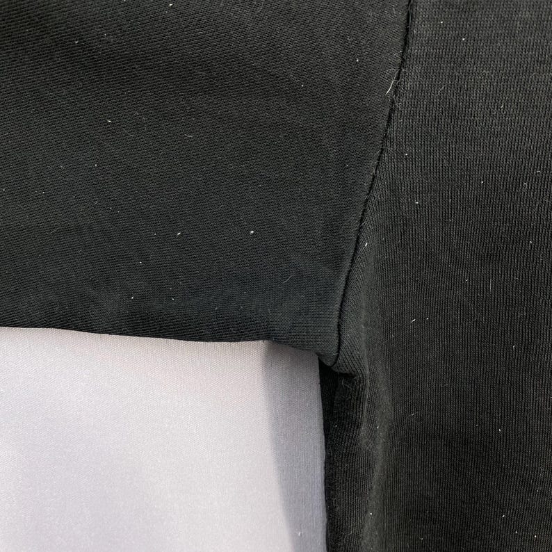 May include: Close-up of a black fabric garment, possibly a shirt or top. The image highlights the texture and stitching details of the material. A white fabric is visible in the lower portion of the image.