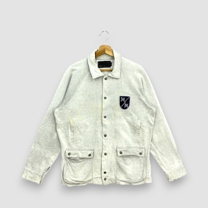 May include: Light gray jacket with a button-up front, two front pockets, and a small patch on the chest. The jacket is hanging on a wooden hanger. The patch features a navy blue emblem.