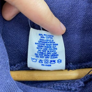 K&ouml;nnte beinhalten: Nahaufnahme eines wei&szlig;en Etiketts mit Pflegehinweisen, das an einem marineblauen Kleidungsst&uuml;ck befestigt ist. Das Etikett enth&auml;lt die Zusammensetzung aus 50 % Polyester und 50 % Baumwolle sowie Wasch- und Trocknungsanweisungen. Die RN-Nummer lautet 61626.