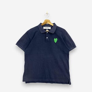 Può includere: Una polo a maniche corte blu navy con colletto, abbottonatura a due bottoni e una toppa verde a forma di cuore con due occhi sul petto sinistro. L'etichetta interna sul collo riporta la scritta PLAY COMME des GARÇONS.