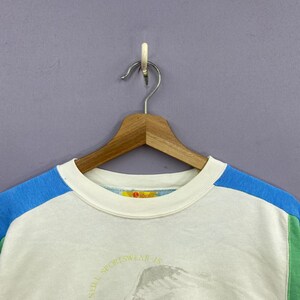 Puede incluir: Sudadera blanca de cuello redondo con mangas de bloques de color azul y verde. La camisa est&aacute; colgada de una percha de madera. El texto "Still Sportswear Is" es visible en la parte inferior de la camisa.