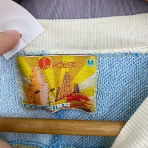 Puede incluir: Primer plano de una sudadera con estampado azul y blanco con un parche bordado amarillo y naranja. El parche presenta el texto "Just Shower" y un dise&ntilde;o de paisaje urbano. La sudadera tiene un cuello blanco y una etiqueta de talla "M".