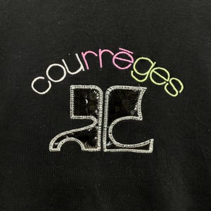 May include: Black garment with the word "courr&egrave;ges" embroidered in white, pink, and green. Below the word is a sequined design with white stitching. The garment is a solid black color.