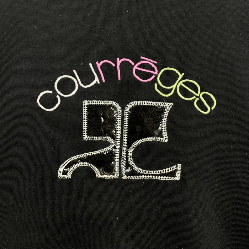 May include: Black garment with the word "courr&egrave;ges" embroidered in white, pink, and green. Below the word is a sequined design with white stitching. The garment is a solid black color.