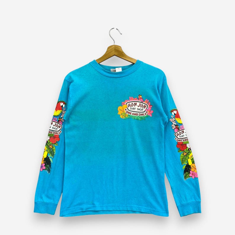 K&ouml;nnte beinhalten: Ein helles, t&uuml;rkisfarbenes Langarmshirt mit einer Grafik auf der Brust und an beiden &Auml;rmeln. Die Brustgrafik zeigt den Text "RON JON SURF SHOP ONE AND ONLY", umgeben von tropischen Blumen und einem Papagei. &Auml;hnliche Blumen- und Papageienmotive befinden sich auf den &Auml;rmeln.