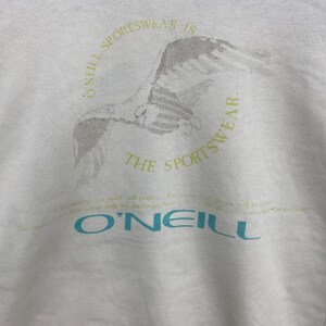 Puede incluir: Sudadera blanca con un gr&aacute;fico de un p&aacute;jaro en vuelo, rodeado por el texto "O'Neill Sportswear is the Sportswear". El nombre de la marca "O'Neill" est&aacute; impreso en azul verdoso. Tambi&eacute;n hay texto adicional en verde claro.