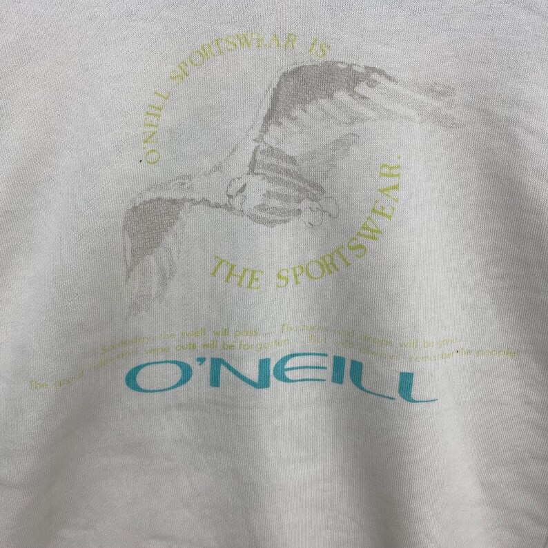 Puede incluir: Sudadera blanca con un gr&aacute;fico de un p&aacute;jaro en vuelo, rodeado por el texto "O'Neill Sportswear is the Sportswear". El nombre de la marca "O'Neill" est&aacute; impreso en azul verdoso. Tambi&eacute;n hay texto adicional en verde claro.