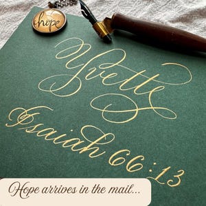 May include: A dark green card featuring gold calligraphy that reads Yvette and Isaiah 66:13. A wooden dip pen with a metal nib rests on the card, accompanied by a small round pendant with a gold rim and the word hope written in black script.