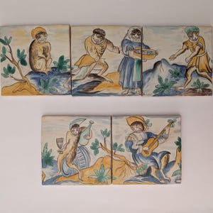 Puede incluir: Conjunto de seis azulejos de cerámica con ilustraciones caprichosas de figuras con vestimenta de época. Los azulejos representan escenas con monos, instrumentos musicales y comida, ambientadas en un paisaje con árboles y montañas. La paleta de colores incluye azules, amarillos y marrones.