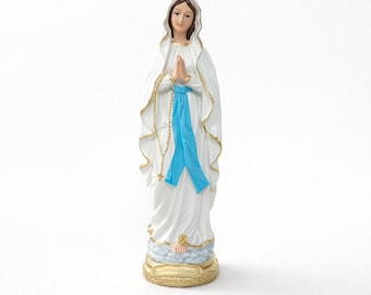 Op de afbeelding: Een wit beeld van een vrouw met een blauwe sjerp en een goudkleurige basis. Het beeld heeft een serene uitdrukking en draagt een witte jurk met gouden randen. Een rozenkrans hangt van haar handen.