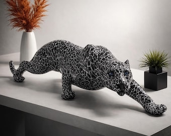 Op de afbeelding: Een zwart-wit decoratief luipaardbeeld met blauwe ogen, in een kruipende houding. Het beeld heeft een gebarsten patroonontwerp. Het luipaard staat op een grijs oppervlak, met een witte vaas en plant op de achtergrond.