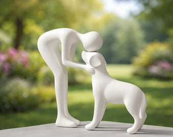 Op de afbeelding: Een witte keramische sculptuur die een persoon afbeeldt die een hond omhelst. De minimalistische figuren zijn glad en gestileerd, waarbij de persoon zich buigt om de kop van de hond aan te raken. De sculptuur staat op een neutrale ondergrond, met een vage groene achtergrond.