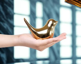 Op de afbeelding: Een goudkleurig decoratief vogelbeeldje met een strak, modern design. De vogel wordt in een hand gehouden en toont zijn gepolijste, reflecterende oppervlak. Het beeldje heeft een lange, gebogen staart en een kleine snavel.
