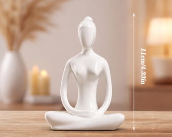 Op de afbeelding: Een witte keramische yoga figuur in een zittende pose. De figuur is 11 cm hoog en 7,5 cm breed. De figuur staat op een houten oppervlak. De achtergrond bevat een vaas met gedroogde planten en kaarsen.