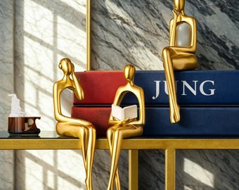 Op de afbeelding: Drie goudkleurige abstracte sculpturen van zittende figuren, een leest een boek, een ander heeft een hand aan het hoofd en de derde aan de kin. De sculpturen staan op een gouden metalen tafel met rode en blauwe boeken op de achtergrond.