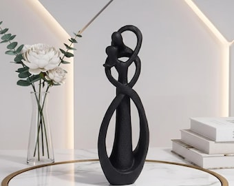 Op de afbeelding: Een zwarte abstracte sculptuur van twee verstrengelde figuren, met een lusvormig ontwerp. De sculptuur staat op een dienblad met marmeren blad en een gouden rand. Een vaas met witte bloemen en groen staat op de achtergrond.