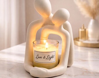 Op de afbeelding: Een witte keramische sculptuur van twee omarmende figuren die een brandende kaars in een glazen pot vasthouden. De kaars heeft een label met de tekst "Love & Light". De sculptuur staat op een marmeren oppervlak.