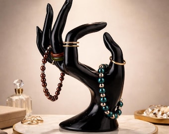 Op de afbeelding: Een zwarte handvormige sieradenstandaard met diverse sieraden. De standaard heeft ringen en armbanden in goud, bruin en blauwgroen. De standaard staat op een witte en gouden cirkelvormige basis, die de sieraden presenteert.